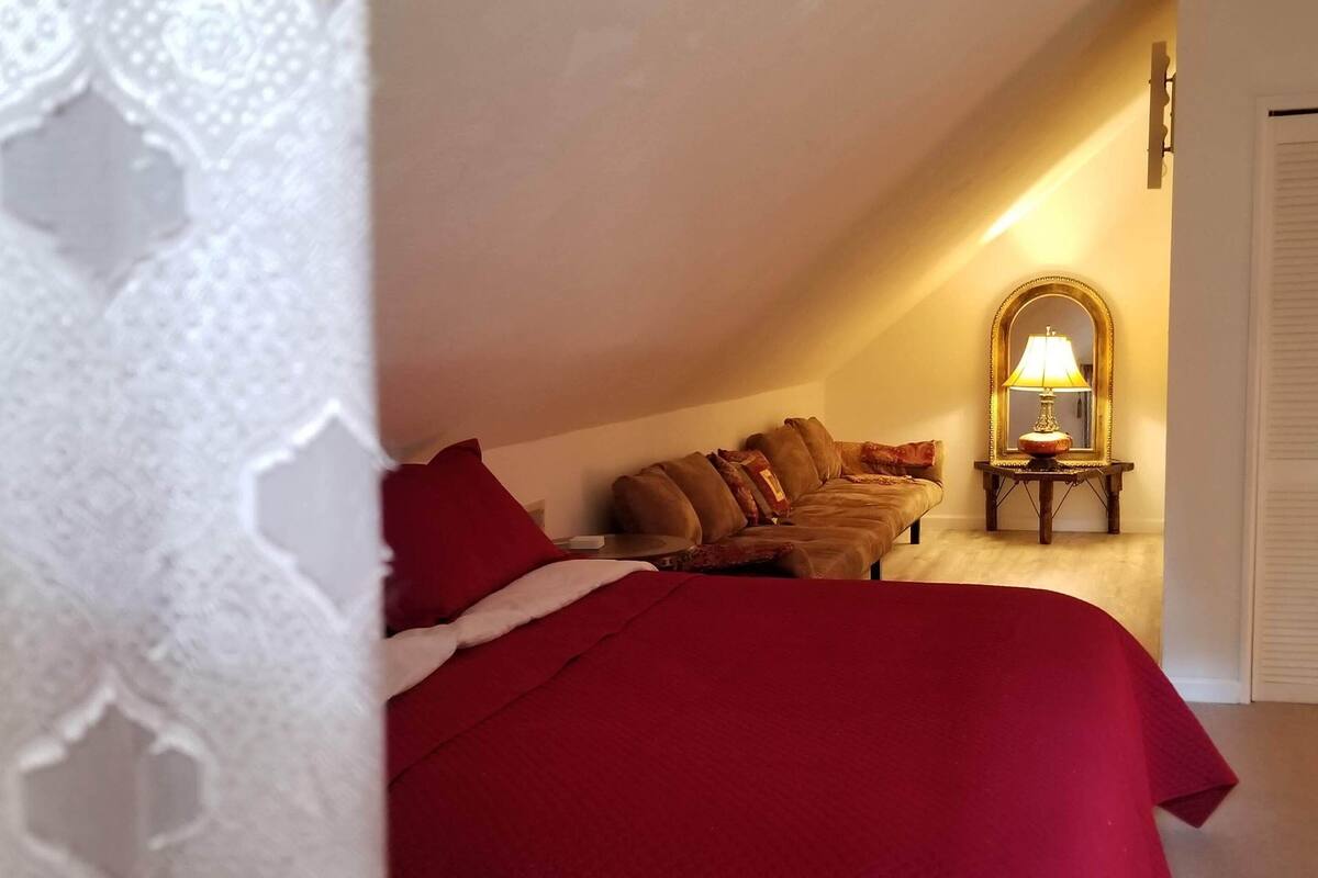 A bedroom with a red bedspread, a wooden nightstand with a lamp, and a mirror on the wall.