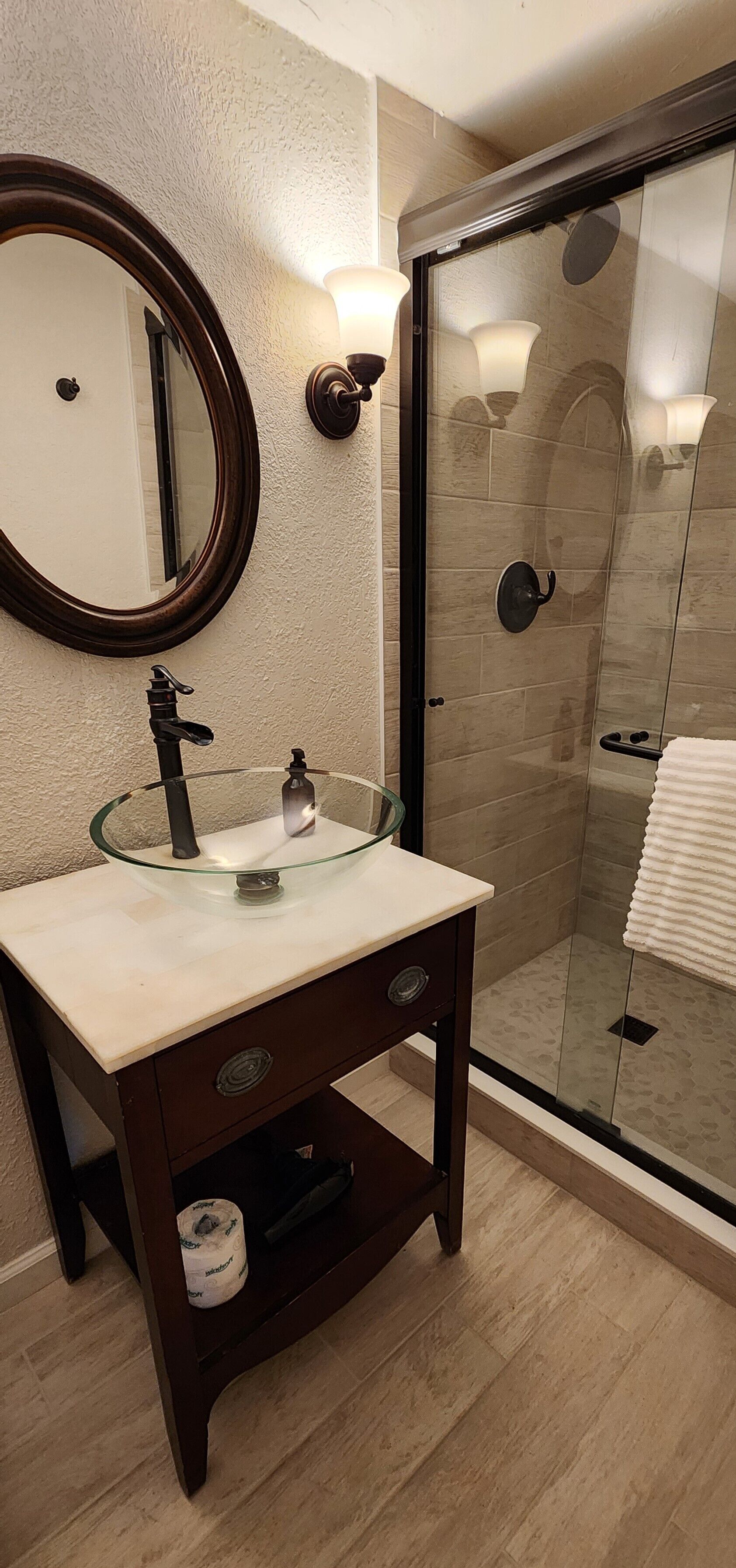 A bathroom with a glass sink, a round mirror, and a glass-enclosed shower.