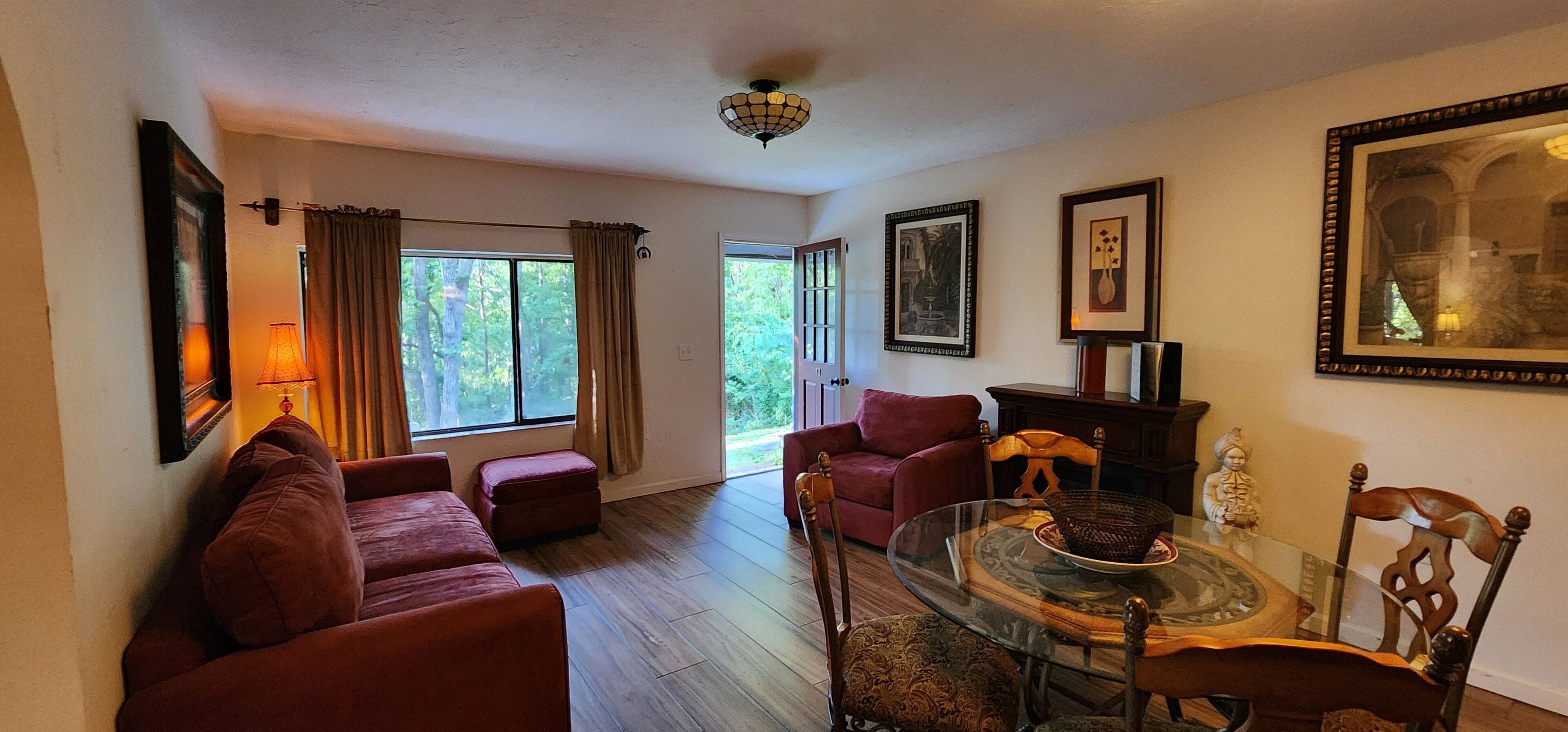 A living room with a glass dining table, red sofas, and a red armchair. There are framed pictures on the walls and a large window with curtains.