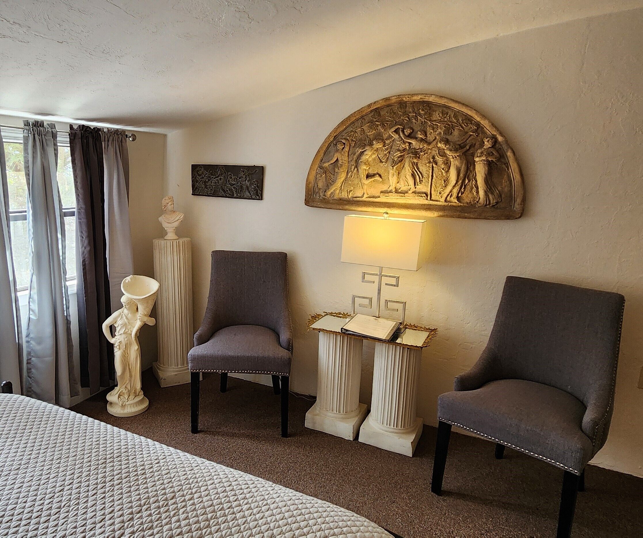 Une chambre d’hôtel comprenant un lit, deux fauteuils, une statue, une lampe fixée au mur et un élément décoratif en relief.