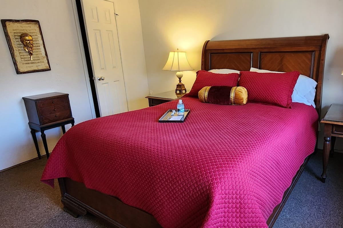 A neatly made bed with a red quilt, two bedside tables with lamps, a small wooden nightstand, and a framed picture on the wall.