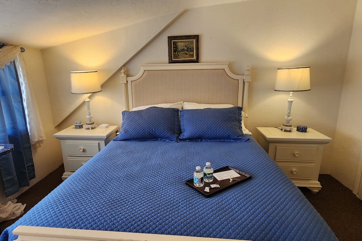A hotel room with a large bed, two bedside tables, a nightstand, a desk, a chair, a window with curtains, and a picture on the wall.