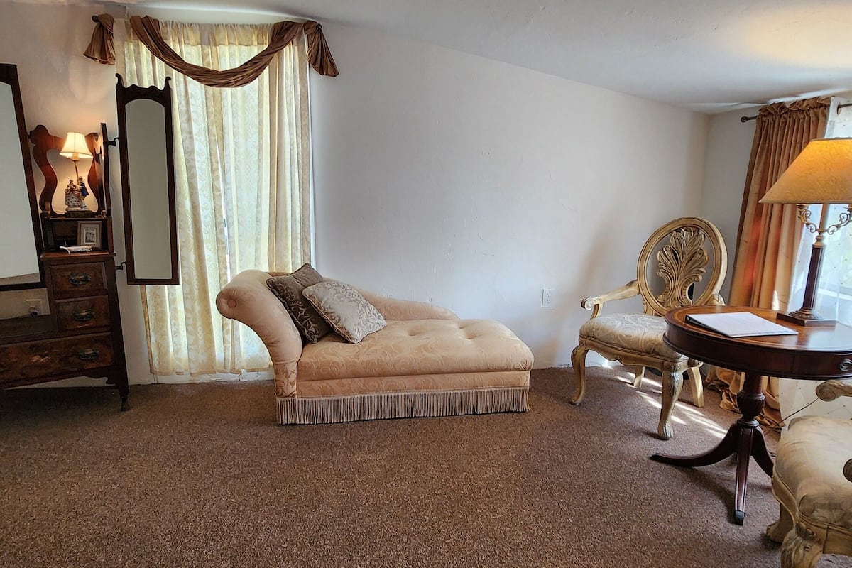 A room with a vintage sofa, a wooden dresser, a mirror, and a table with a lamp.