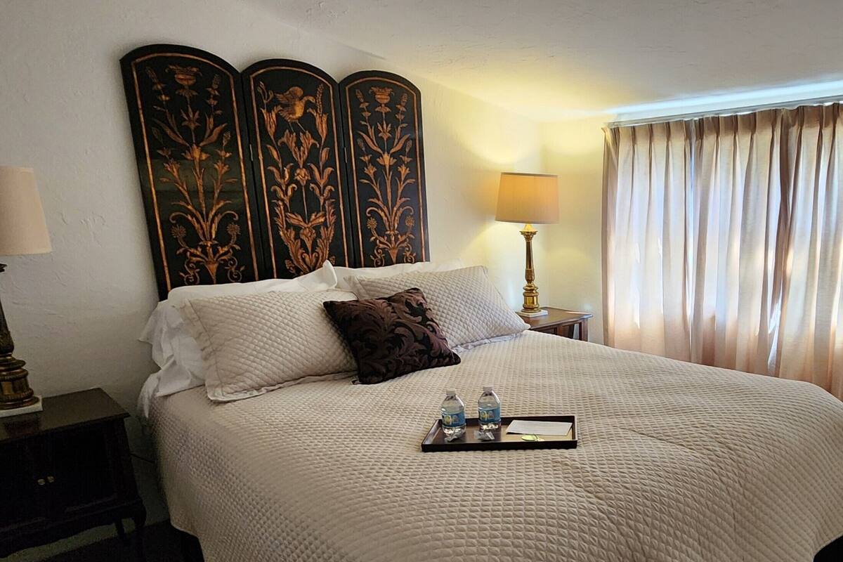 A hotel room with a large bed, a nightstand with a lamp, a tray with water bottles, and a decorative headboard.