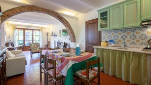 Un comedor con una mesa preparada para cuatro, un sillón floral y una cocina con armarios verdes y un salpicadero de azulejos.