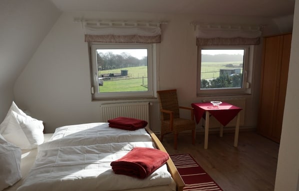 Ein Schlafzimmer mit zwei Betten, einem Sessel, einem Tisch und zwei Fenstern mit Blick auf ein grünes Feld.
