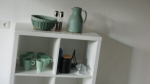 Un área de cocina con un armario blanco que contiene tazas verdes y una tetera verde. Encima del armario, hay tres cuadros enmarcados en la pared.
