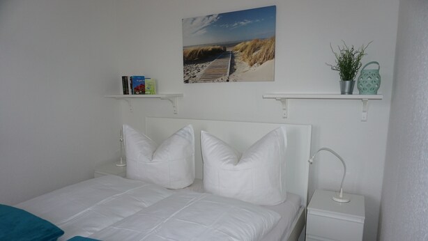 Un dormitorio ordenado con cama blanca, almohadas, una mesita de noche con lámpara, un estante con libros y una planta, y un adorno de pared con temática marina.