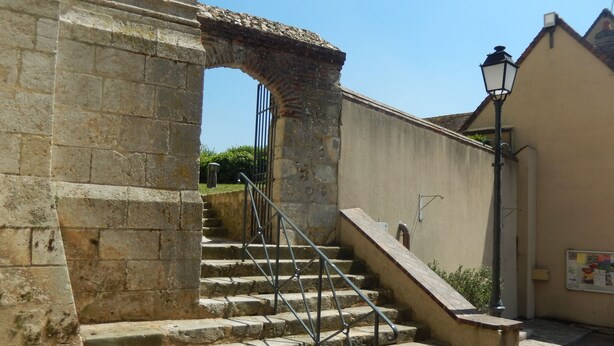 Una escalera de piedra con barandales de metal que lleva a una reja, rodeada de muros de piedra y un farol.