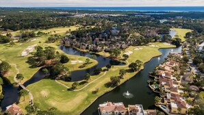 Vista aérea de un campo de golf con elementos acuáticos, rodeado de zonas residenciales y una lejana silueta urbana.