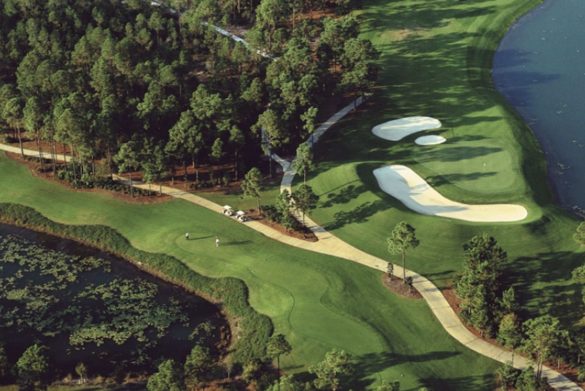 Luftaufnahme eines Golfplatzes mit Sandbunkern, einem Wasserhindernis und bewaldeten Flächen.