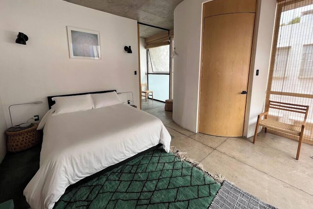 A modern bedroom with a bed, a small bedside table, a green patterned rug, a wooden door, and a view of the outside.