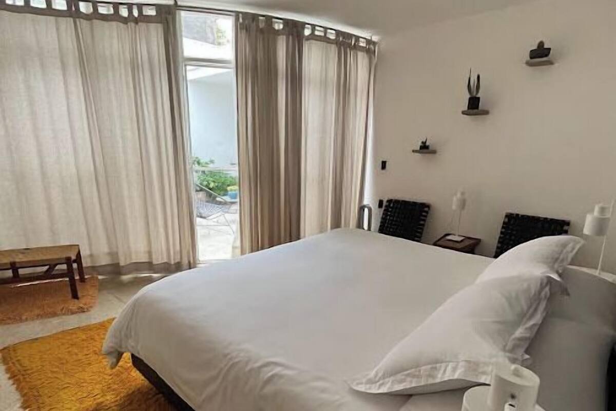 A bedroom with a large bed, a bedside table, a lamp, and a view of the outdoors through a window with curtains.