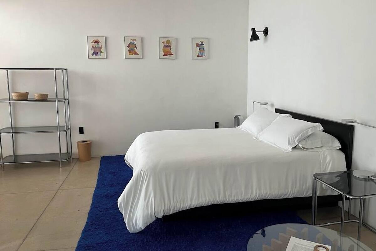 A bedroom with a bed, a blue rug, a metal shelf unit, and four framed pictures on the wall.