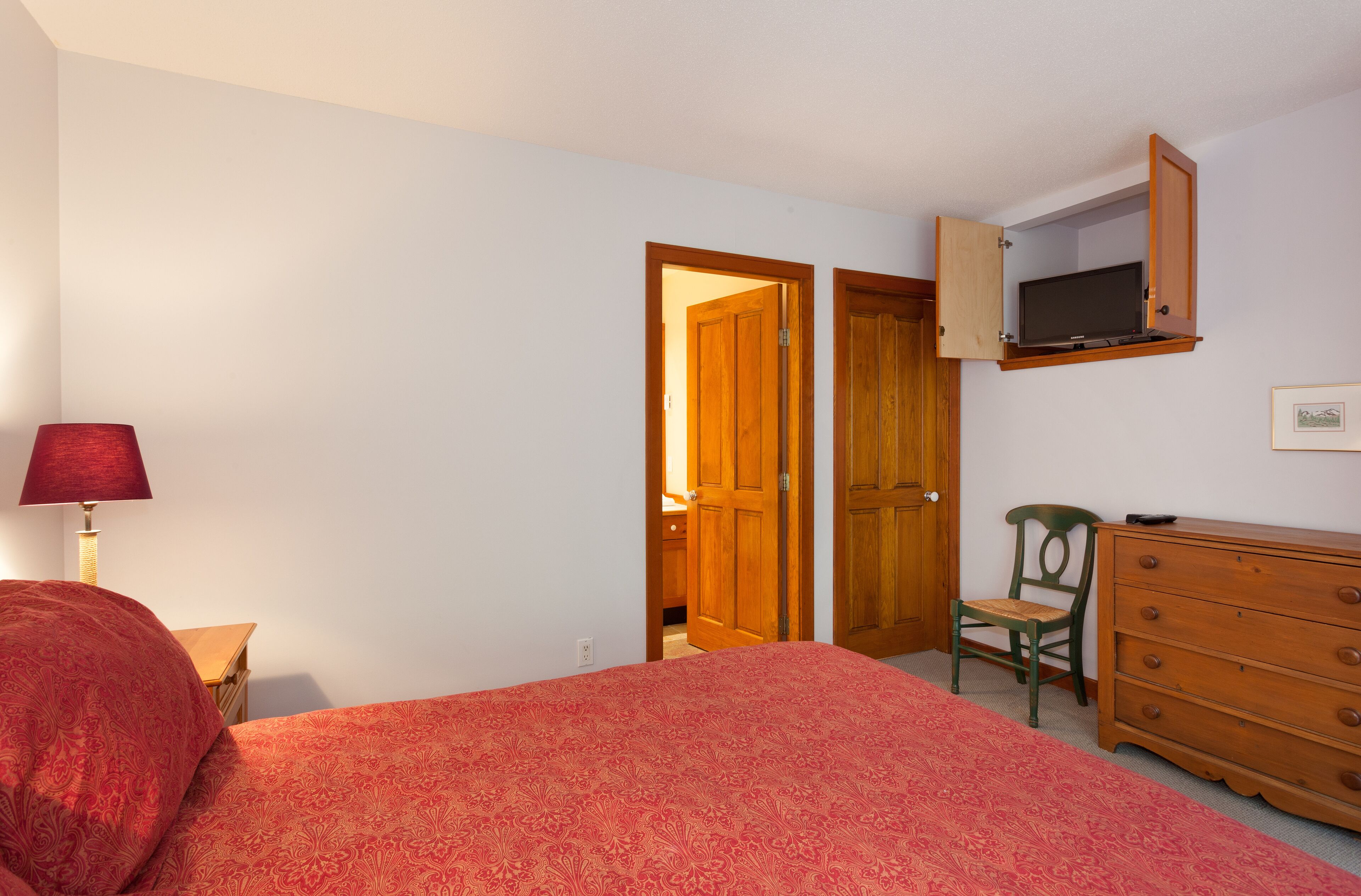 A bedroom with a bed, dresser, TV shelf, lamp, and two doors.