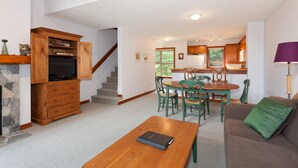 A spacious living area with a wooden coffee table, a green sofa, and a dining table with chairs. A kitchen is visible in the background with wooden cabinets and a window. - Tree Line by Whistler Premier (Whistler)