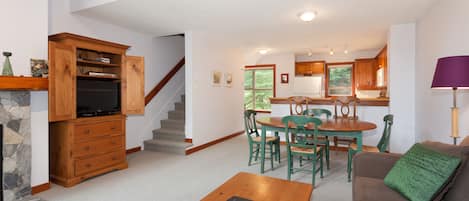 A spacious living area with a wooden coffee table, a green sofa, and a dining table with chairs. A kitchen is visible in the background with wooden cabinets and a window.