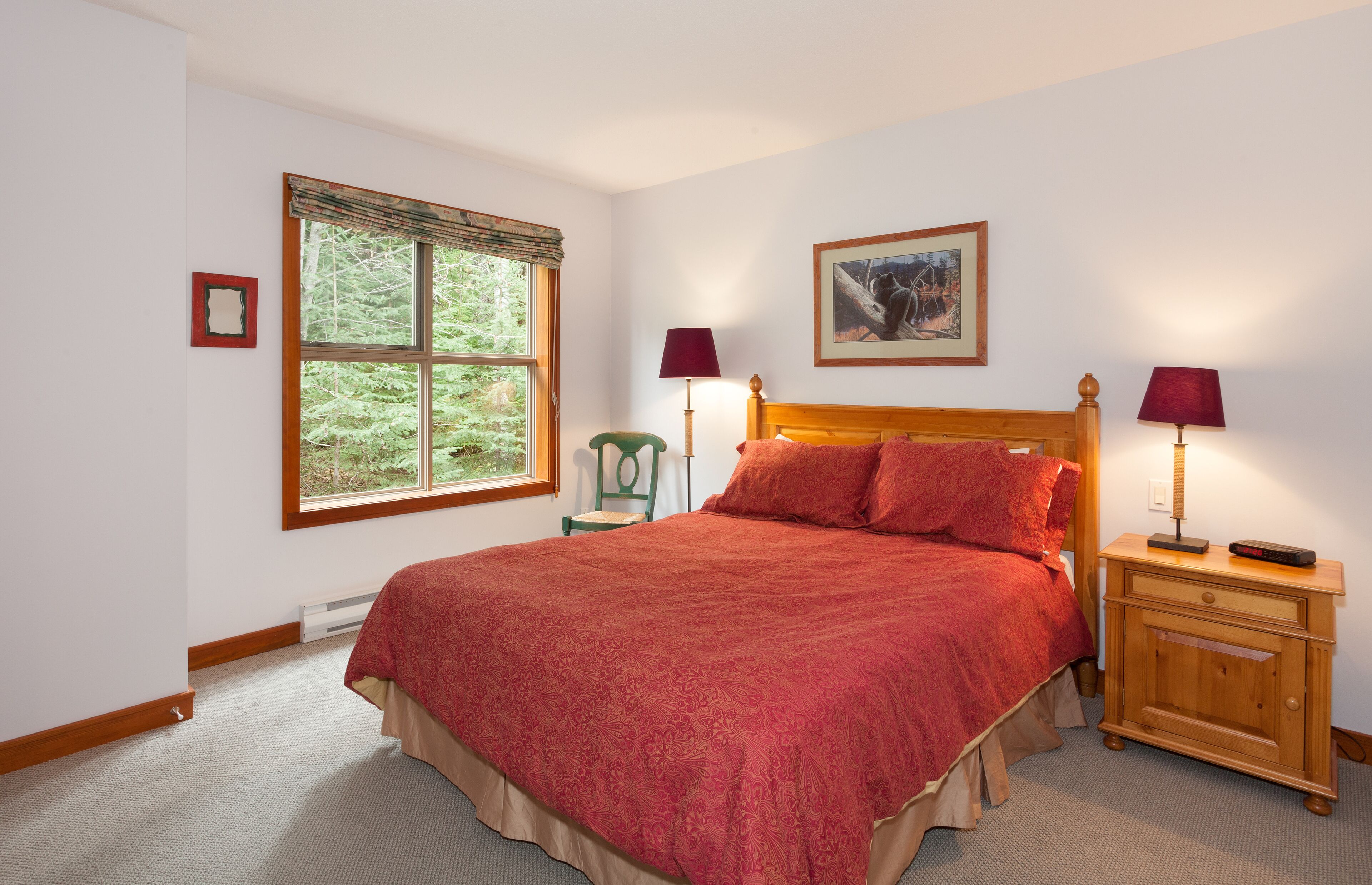 A bedroom with a large bed, two bedside tables with lamps, a window with a view of trees, and a framed picture on the wall.