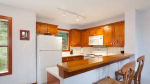 A kitchen with wooden cabinets, a white refrigerator, a microwave, and a wooden bar counter with two chairs. - Tree Line by Whistler Premier (Whistler)