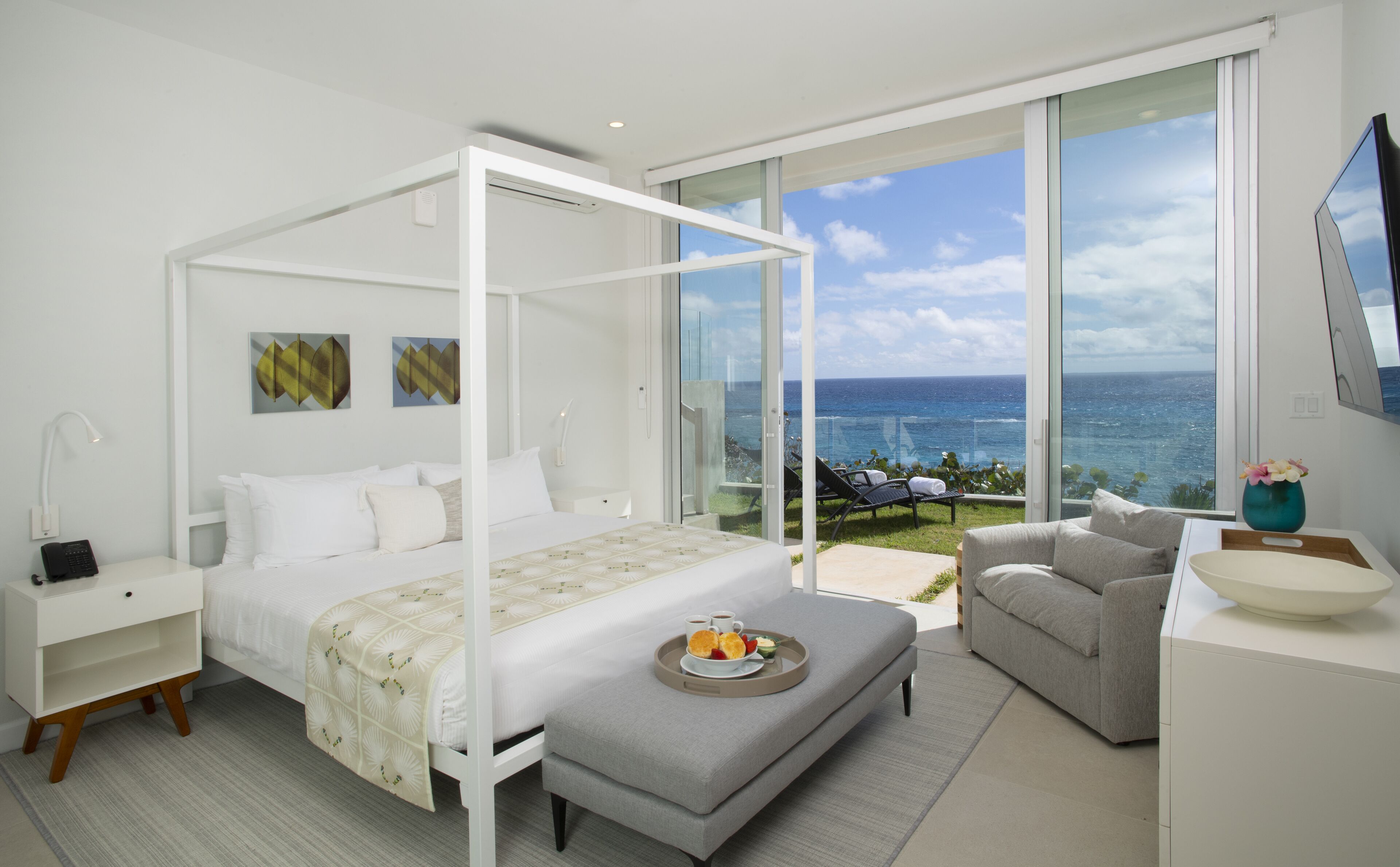 A modern bedroom with a four-poster bed, a sitting area with a view of the ocean, and a dining table with breakfast items.