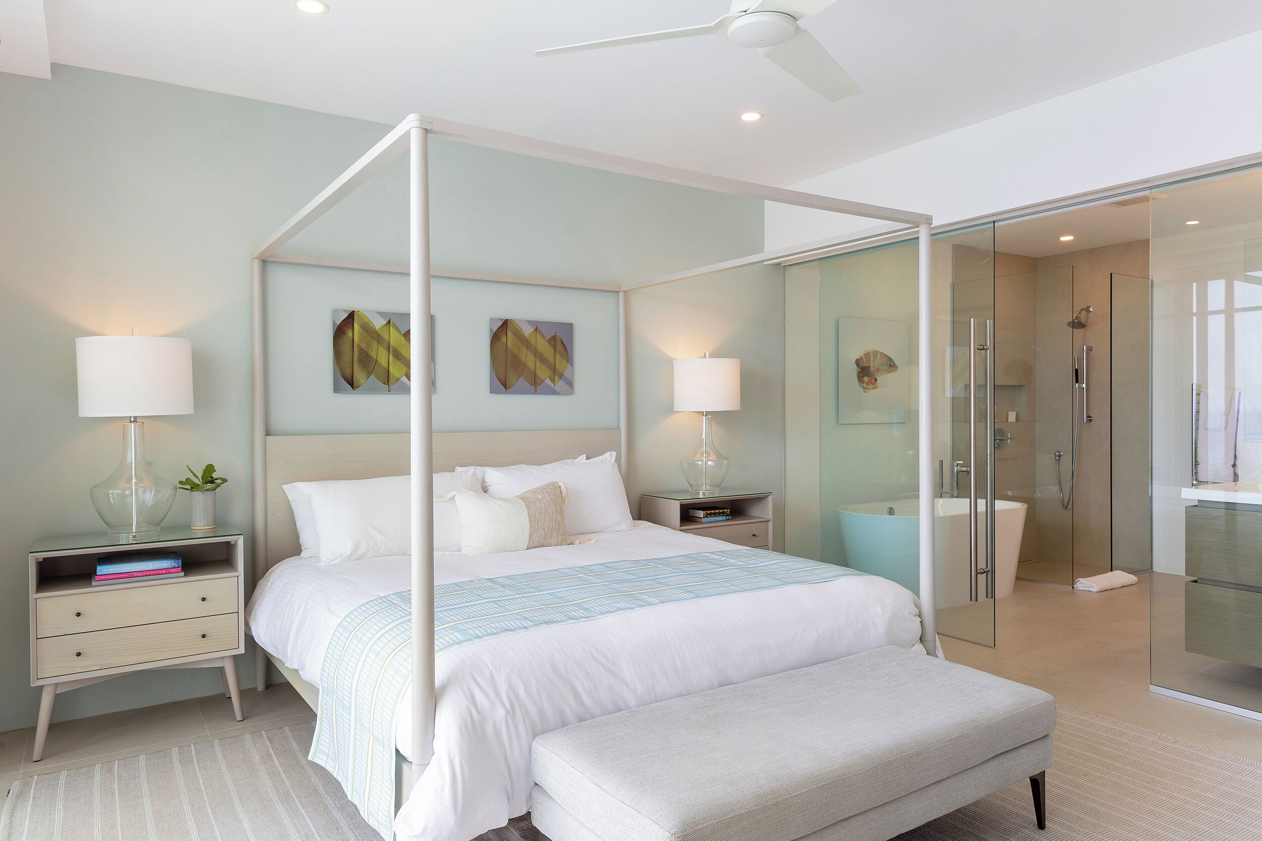 A modern bedroom with a four-poster bed, bedside tables with lamps, a bench, and a glass-enclosed shower area.