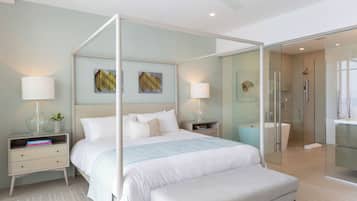 Un dormitorio moderno con cama con dosel, mesitas de noche con lámparas, un banco y una zona de ducha con paredes de vidrio.