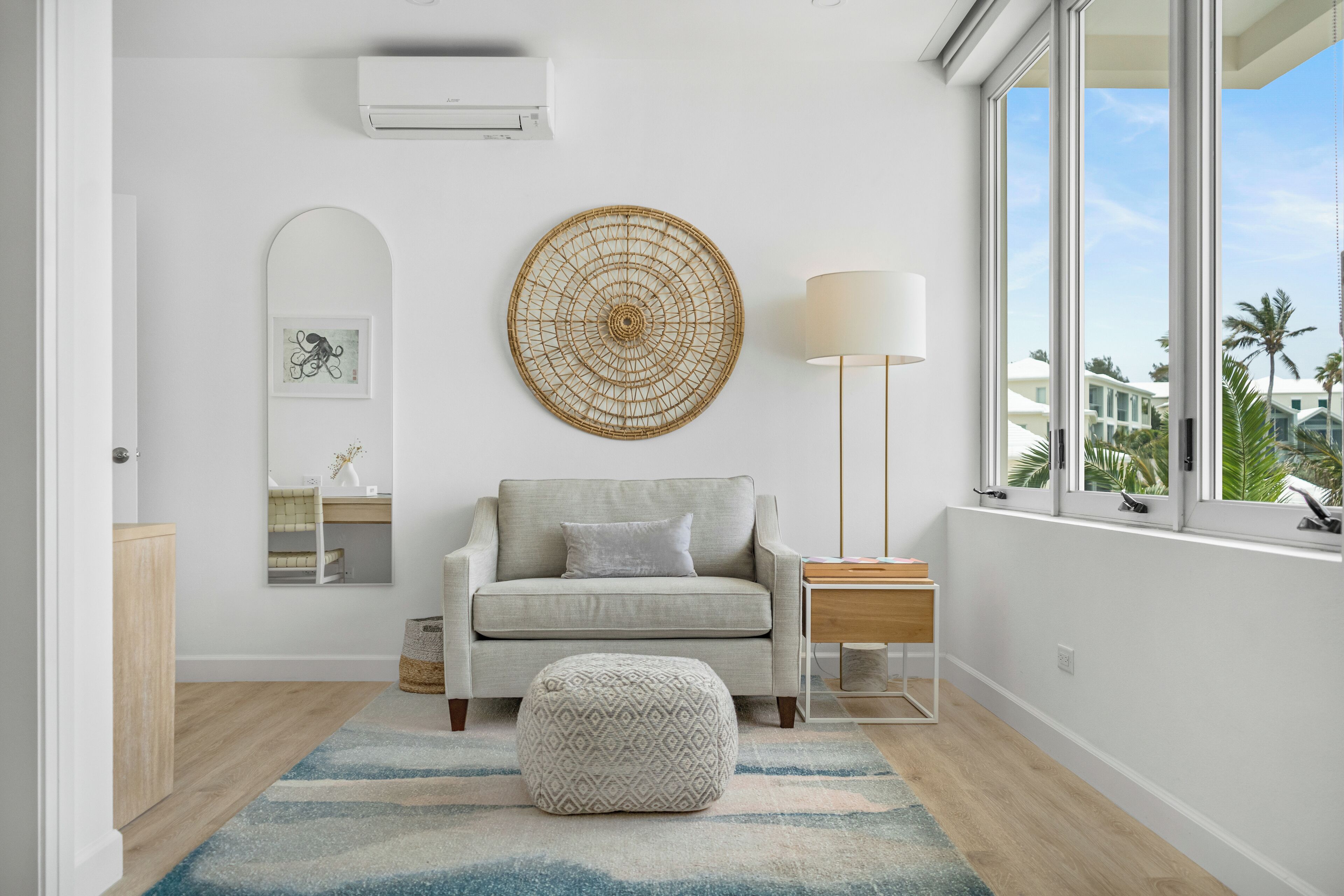 A modern living room with a grey sofa, a round woven wall decoration, a side table with a lamp, and a view of palm trees through large windows.