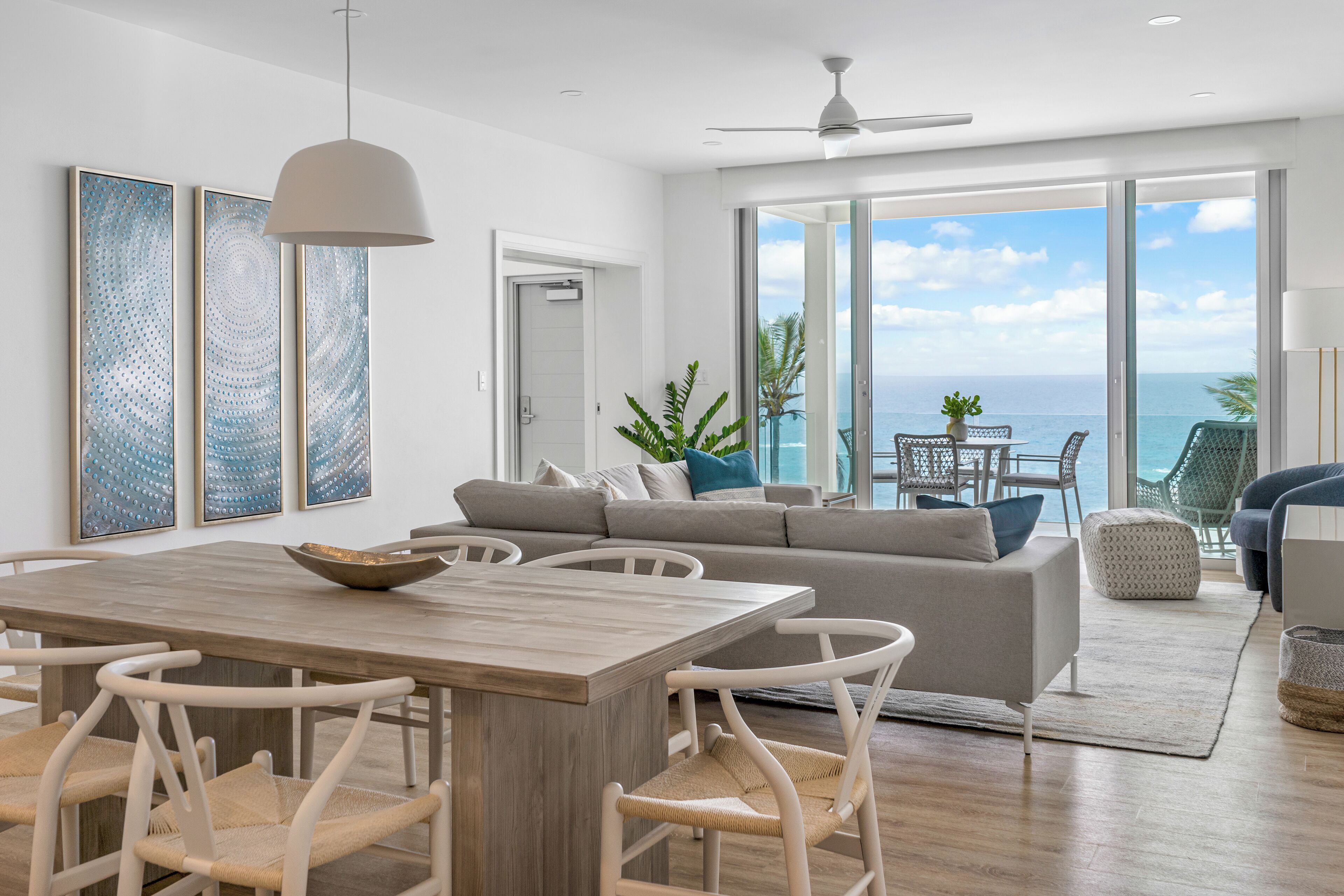 Un salon moderne avec une table à manger en bois, des chaises blanches, un canapé gris et une vue sur l’océan grâce à de grandes portes coulissantes.