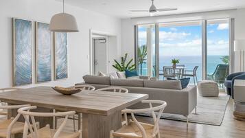 Un salon moderne avec une table à manger en bois, des chaises blanches, un canapé gris et une vue sur l’océan grâce à de grandes portes coulissantes.