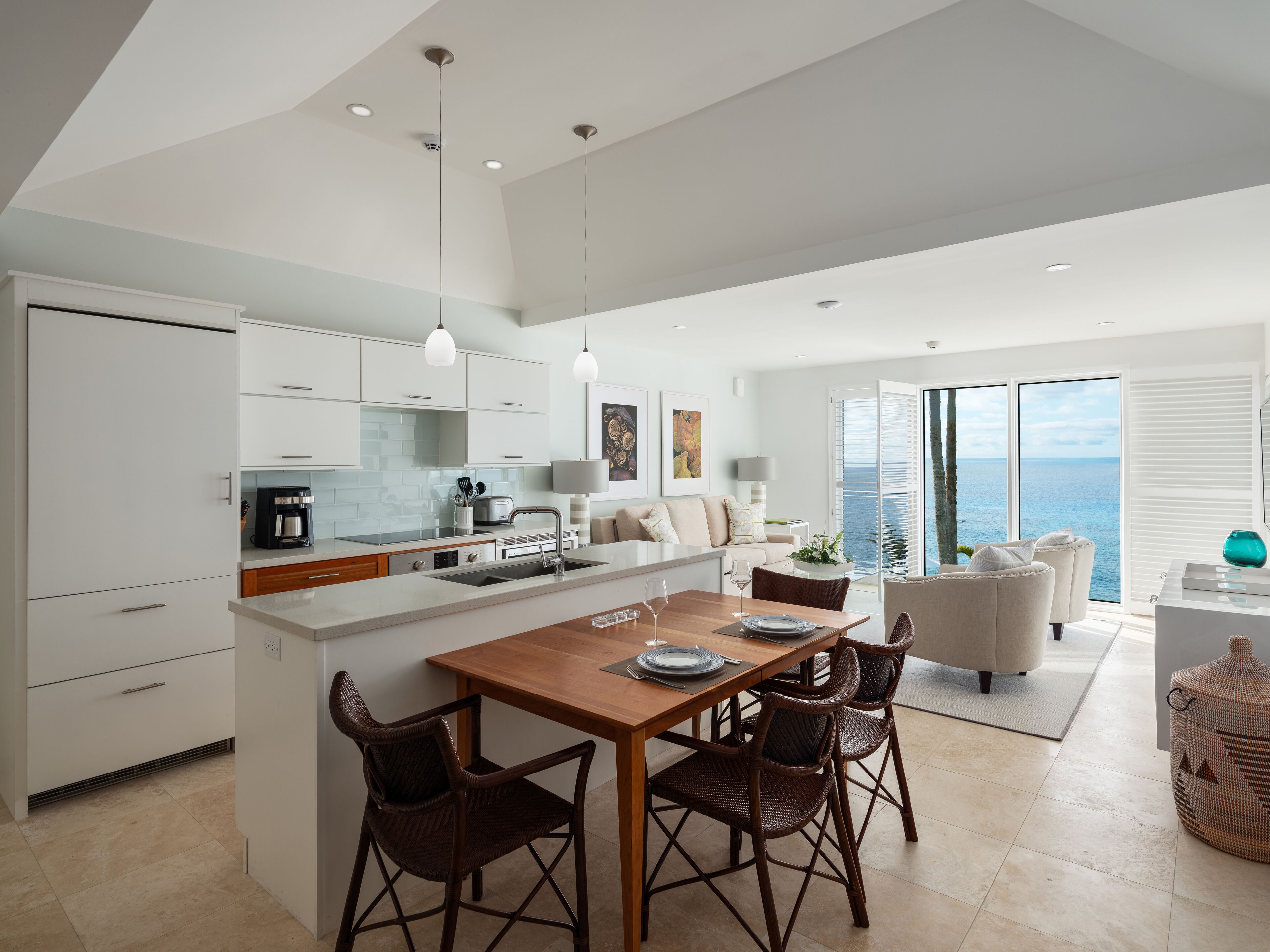 A modern kitchen with white cabinetry, a wooden dining table with four chairs, and a view of the sea through large windows.