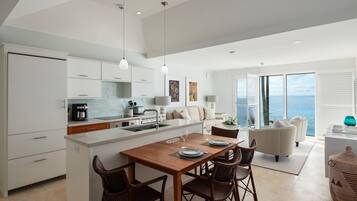 A modern kitchen with white cabinetry, a wooden dining table with four chairs, and a view of the sea through large windows.