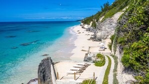 Une plage aux eaux turquoise et limpides, au sable blanc, bordée d’une falaise rocheuse. Des chaises longues et des parasols sont disposés le long du rivage.