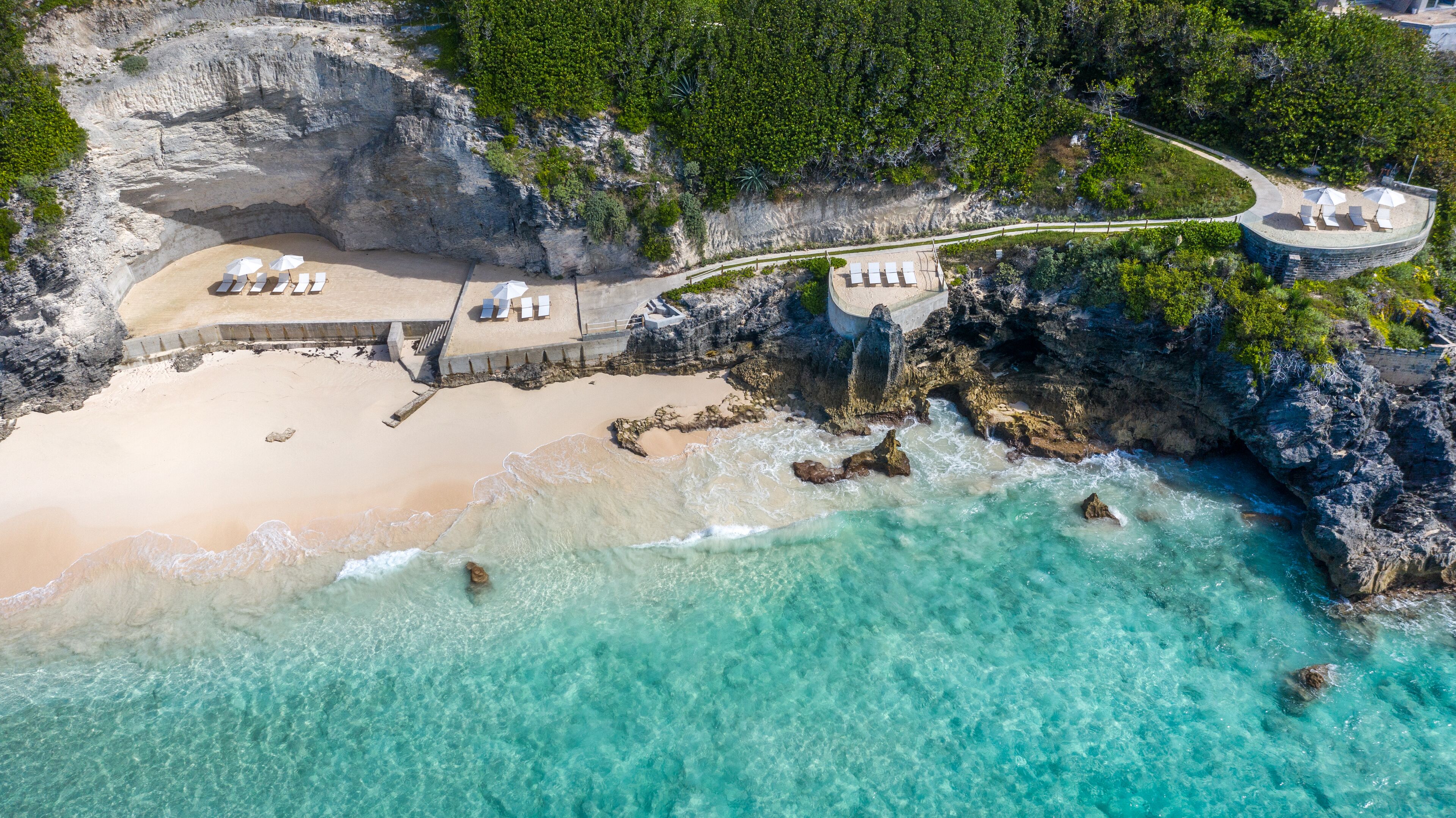 An aerial view of a beach resort with a sandy beach, clear turquoise waters, rocky cliffs, and a pathway leading to a lounge area with sun loungers.