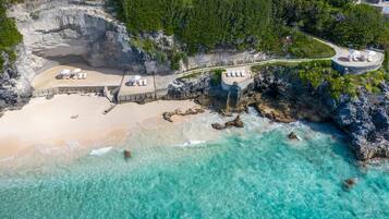 An aerial view of a beach resort with a sandy beach, clear turquoise waters, rocky cliffs, and a pathway leading to a lounge area with sun loungers.