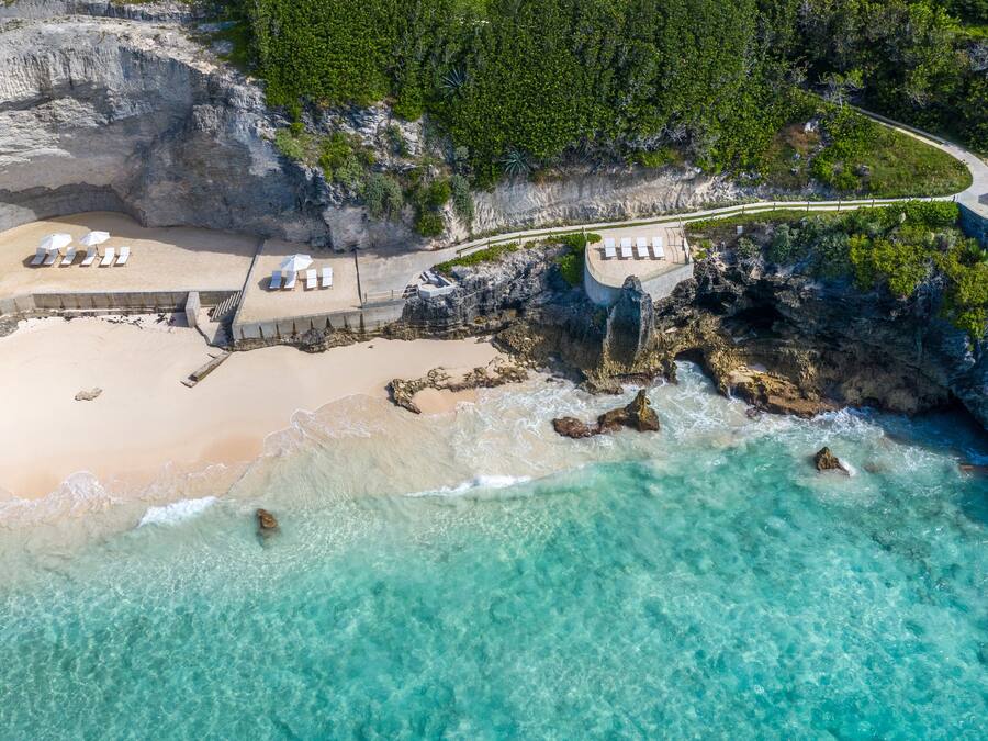An aerial view of a beach resort with a sandy beach, clear turquoise waters, rocky cliffs, and a pathway leading to a lounge area with sun loungers.