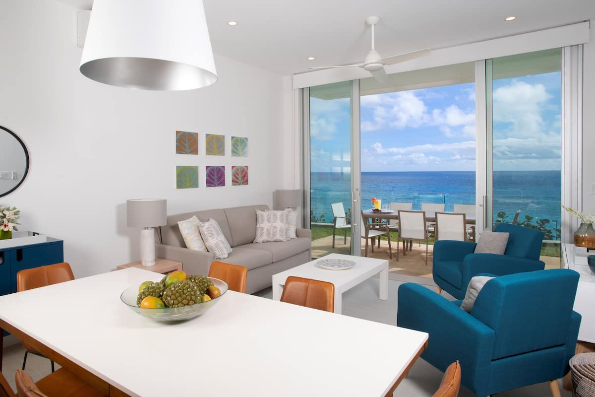 Uma sala de estar moderna com uma janela ampla com vista para o mar, uma área de jantar com uma fruteira e um espaço de estar acolhedor.