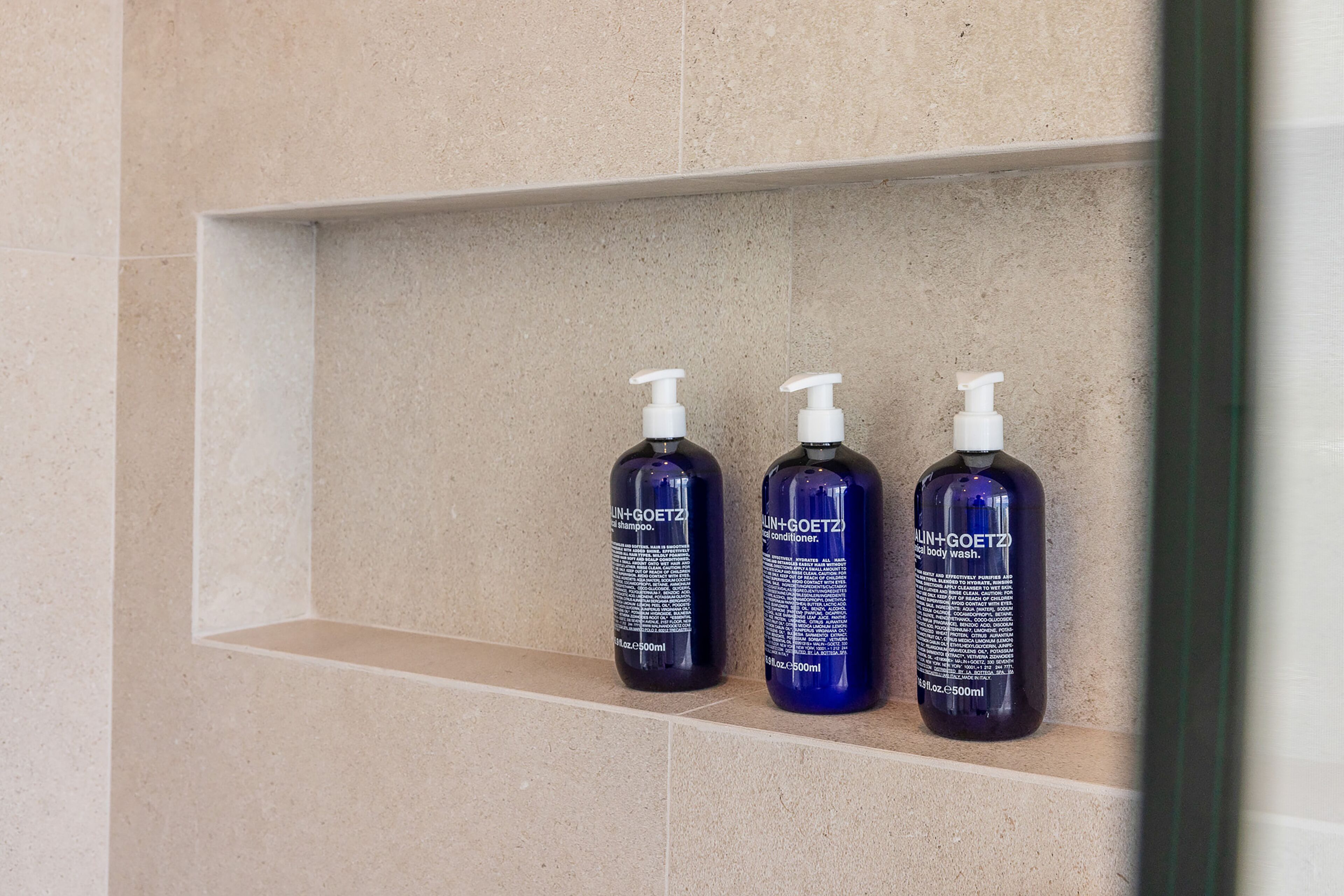 Three bottles of SUN+GOETZ body wash on a beige tiled shelf.