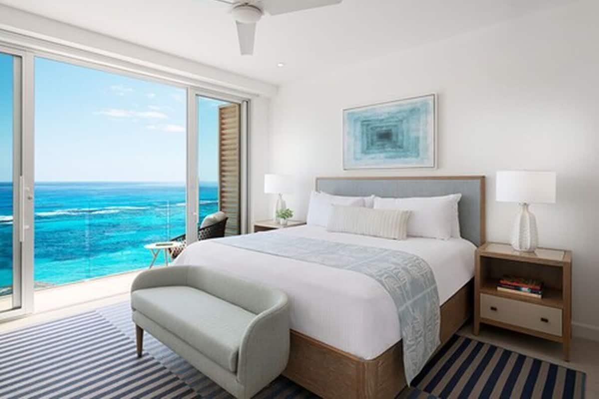 Quarto de hotel moderno com uma cama grande, vista para o oceano, ventoinha no teto e mesa de cabeceira com abajur.