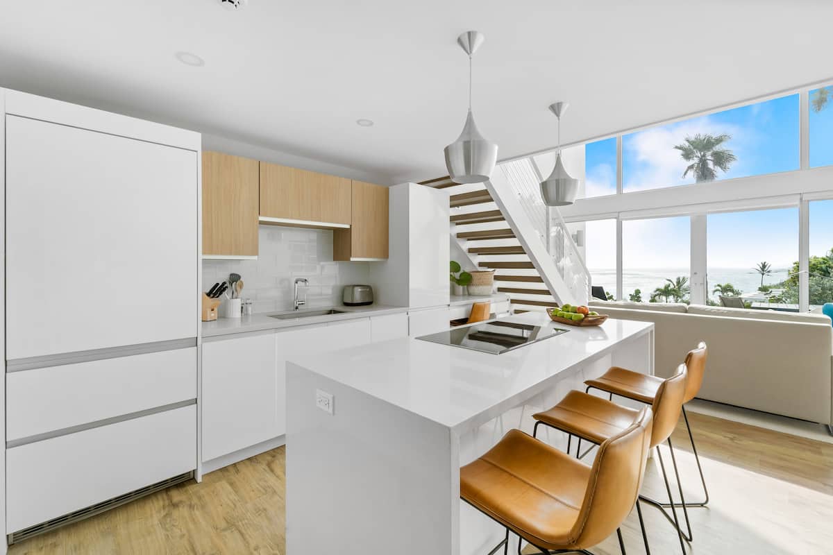 Eine moderne Küche mit weißen Schränken, einer zentralen Kochinsel mit Barhocker und einer Treppe, die zu einem Balkon führt, mit Blick auf den Ozean.