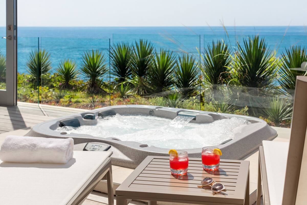 Uma banheira de hidromassagem com vista desimpedida para o oceano, uma toalha e duas bebidas sobre uma mesa.