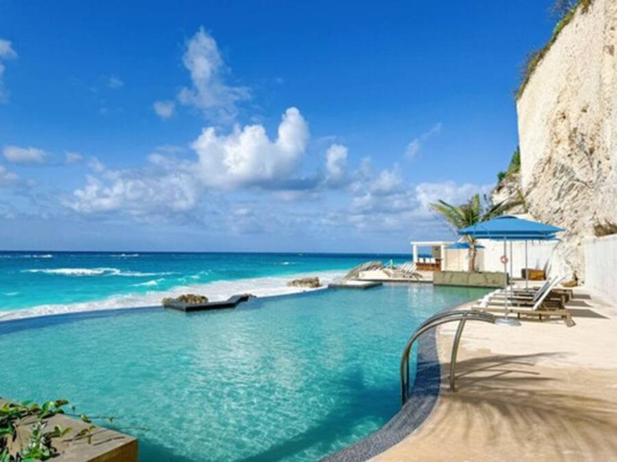 A clear blue pool with a view of the ocean, a small boat, and a white building with a blue roof.