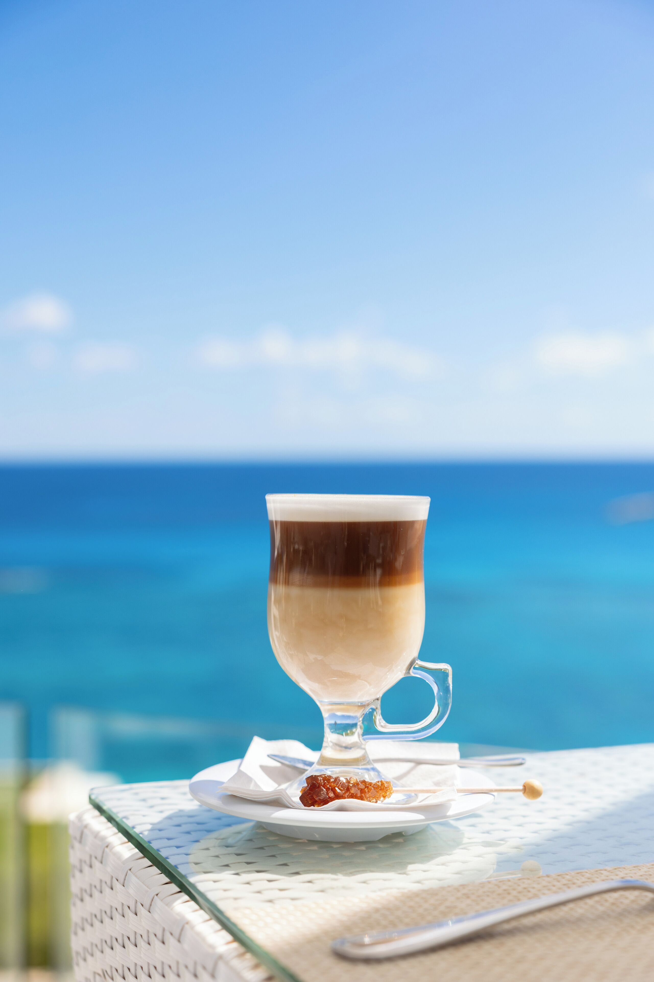 Una taza de vidrio transparente con una bebida en capas sobre un mantel blanco, un plato pequeño con una galleta y una cuchara, frente a un cielo azul y el océano.