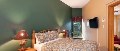 A bedroom with a bed, a nightstand with a lamp, a window with curtains, and a television on the wall.