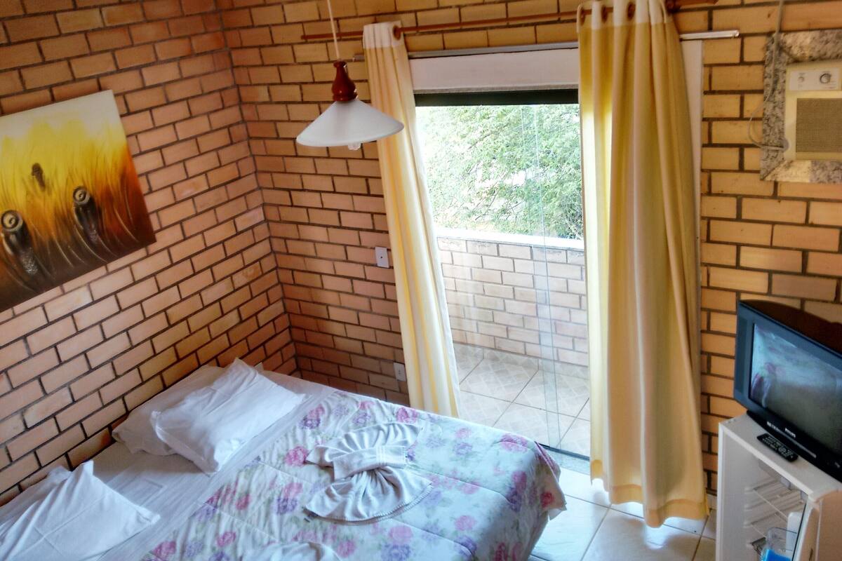 Ein Schlafzimmer mit Backsteinwand, einem Bett mit floralem Bettbezug, einer Deckenleuchte, einem Fernseher und einem Fenster mit Vorhängen.