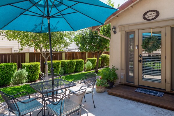 An outdoor patio with a table and chairs, a blue umbrella, and a door with a sign reading "Casa de Mimosas".