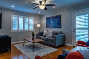 A living room with a gray sofa, a coffee table, and a ceiling fan. There is a window with blinds, a map on the wall, and a door with frosted glass panels.