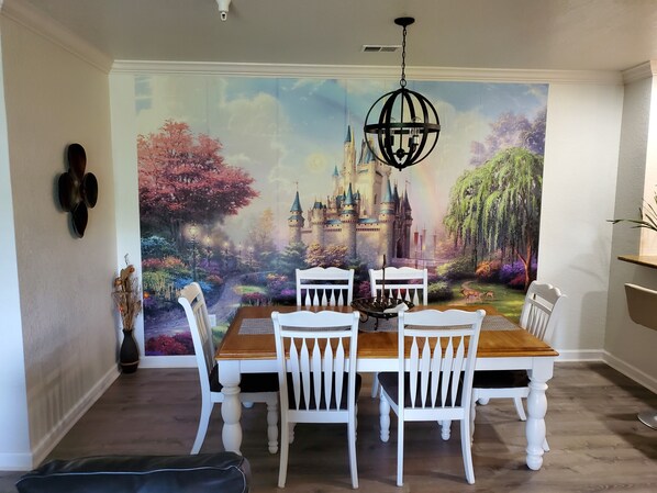 Un comedor con un mural de castillo, sillas blancas, una mesa de madera y una araña negra.