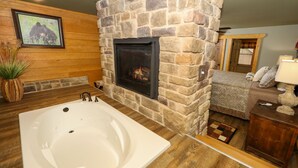 Ein Badezimmer mit Kamin aus Stein, einer großen Badewanne, Holzboden und einer gemütlichen Sitzecke mit Lampe und Kissen.