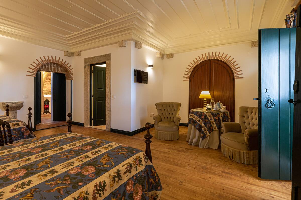 Quarto com uma cama grande, duas poltronas, um armário de madeira e uma mesinha de cabeceira com abajur.