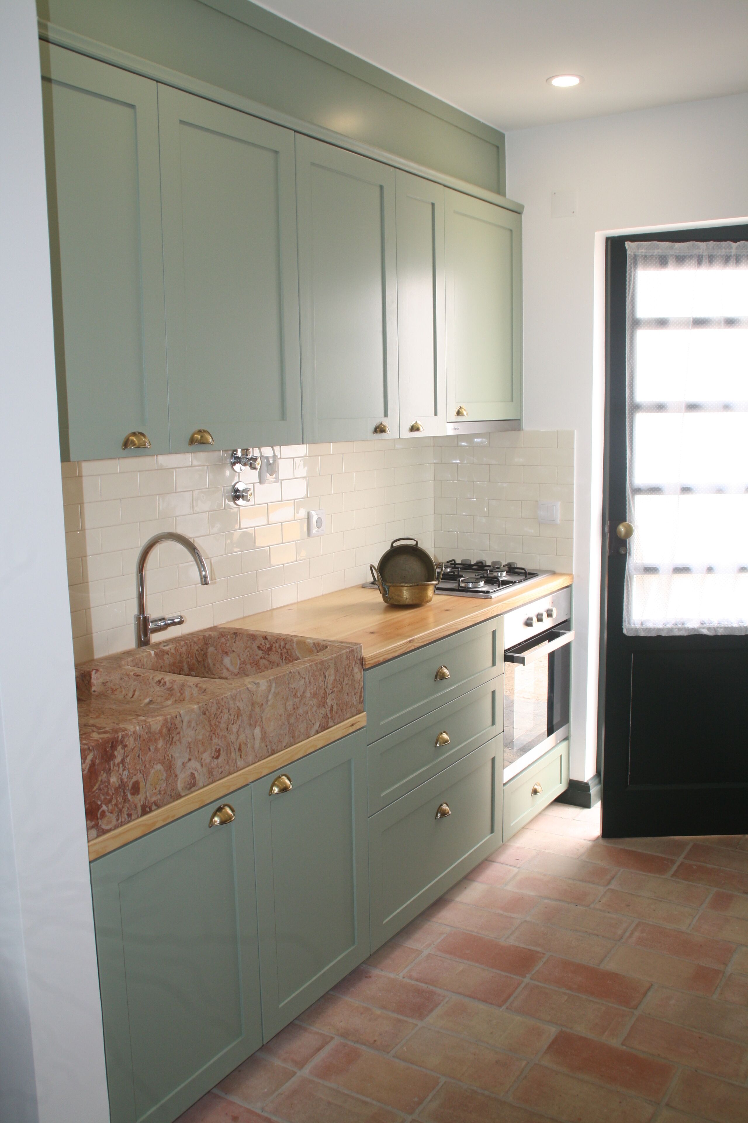 A kitchen with light green cabinets, a marble sink, a stove, and a window with a curtain.
